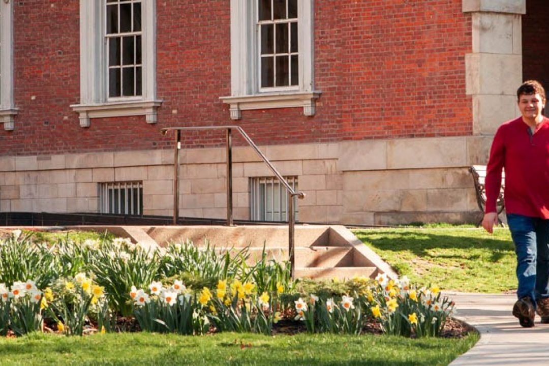 A young man and his black Lab guide dog walking in a park, surrounded by green trees, yellow flowers, and a brick building in the background.