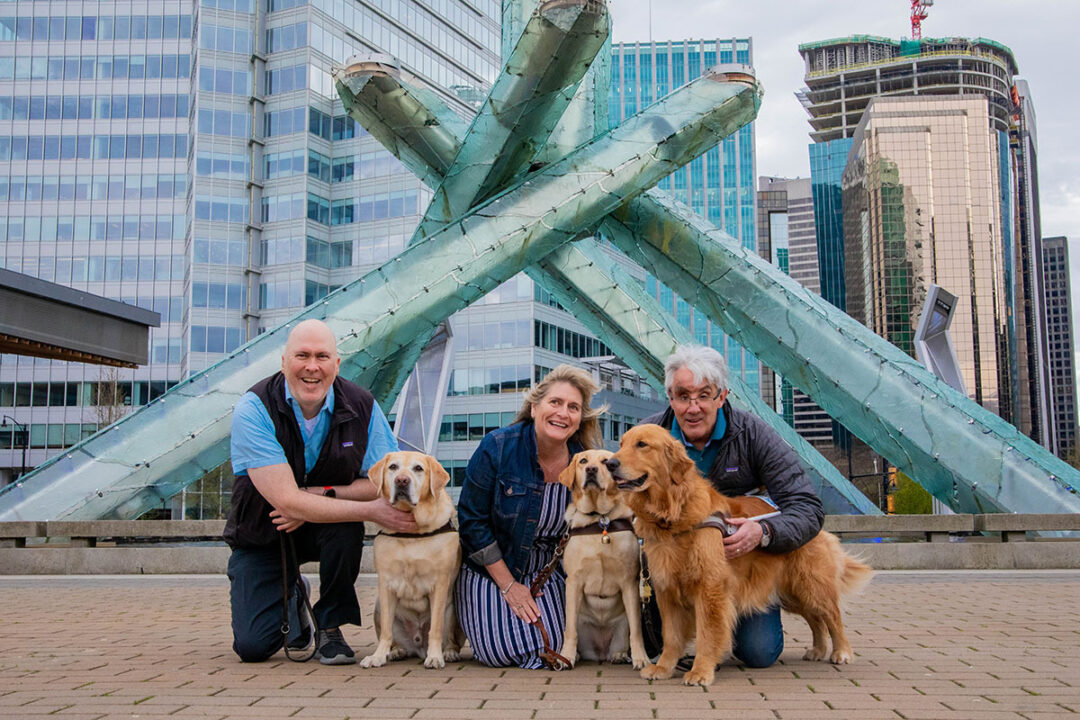 Three people kneel beside their guide dogs in front of a large sculpture in a city plaza.