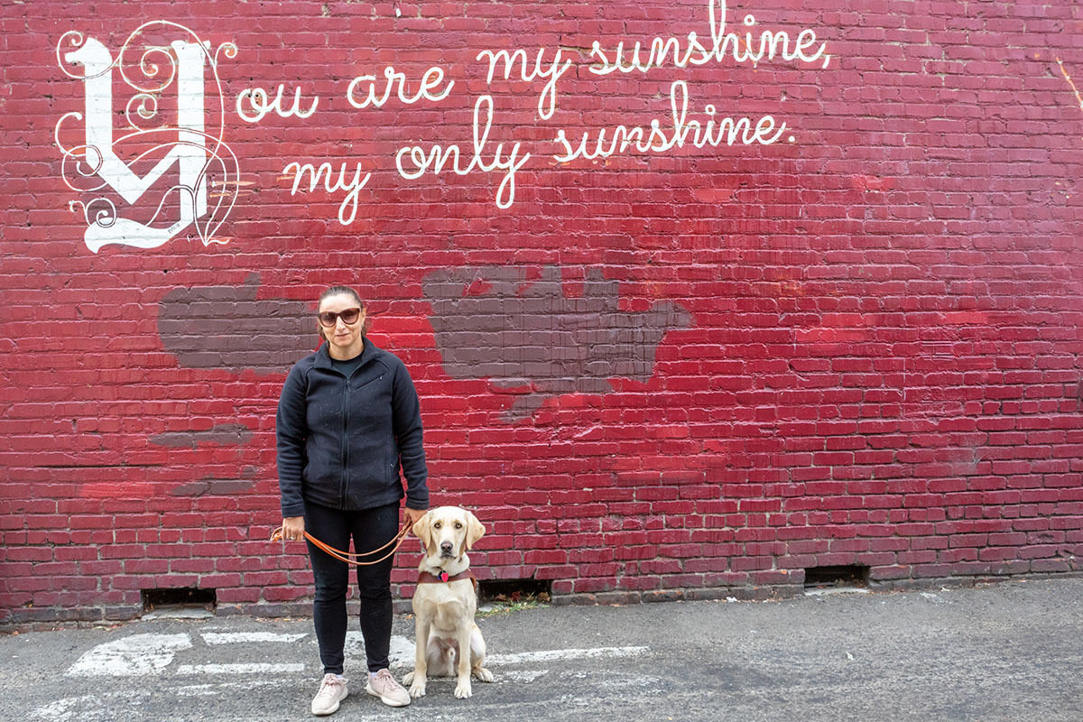A woman and her guide dog stand against a red brick wall painted with the words "You are my sunshine, my only sunshine."