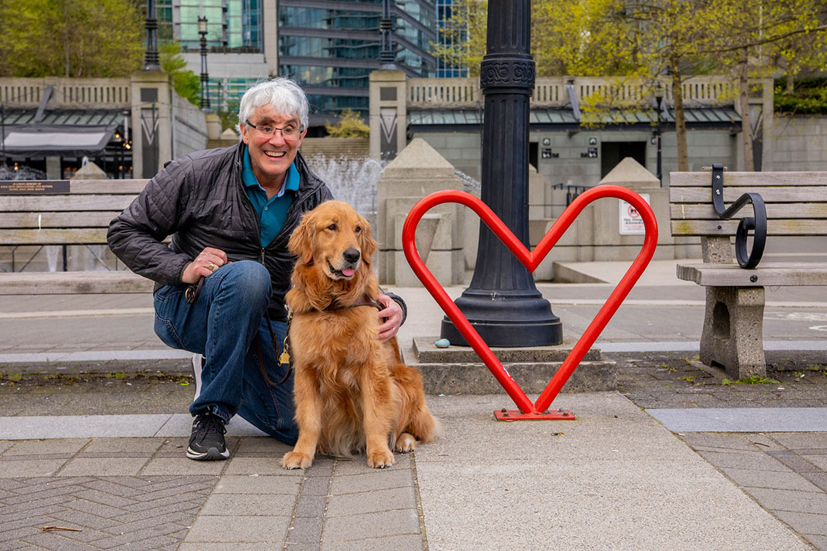 A person kneels next to their guide dog on a city sidewalk near a sculpture in the shape of a heart.