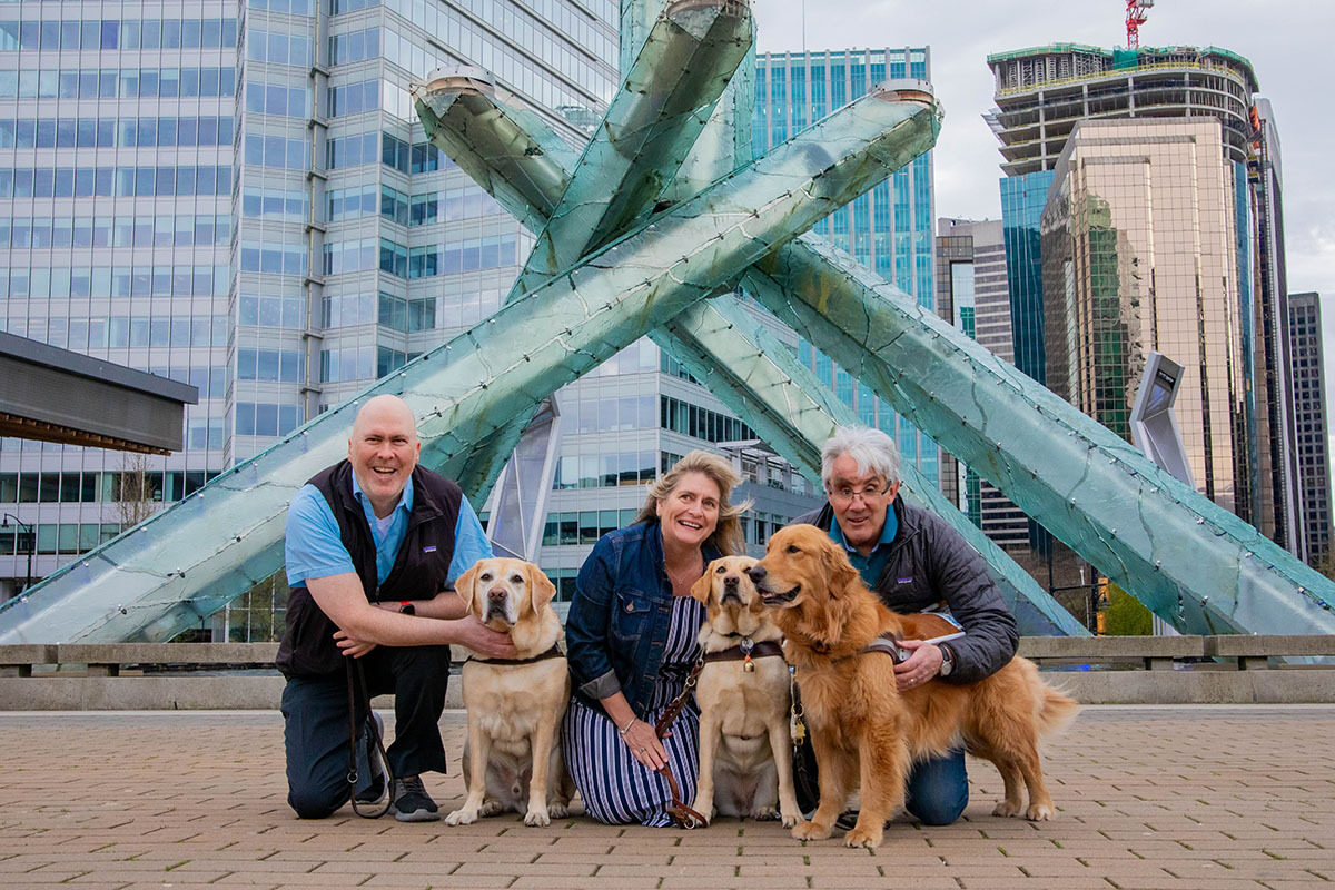 Three people kneel beside their guide dogs in front of a large sculpture in a city plaza.