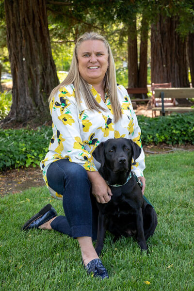 A woman kneels in the grass next to a black Lab guide dog puppy.