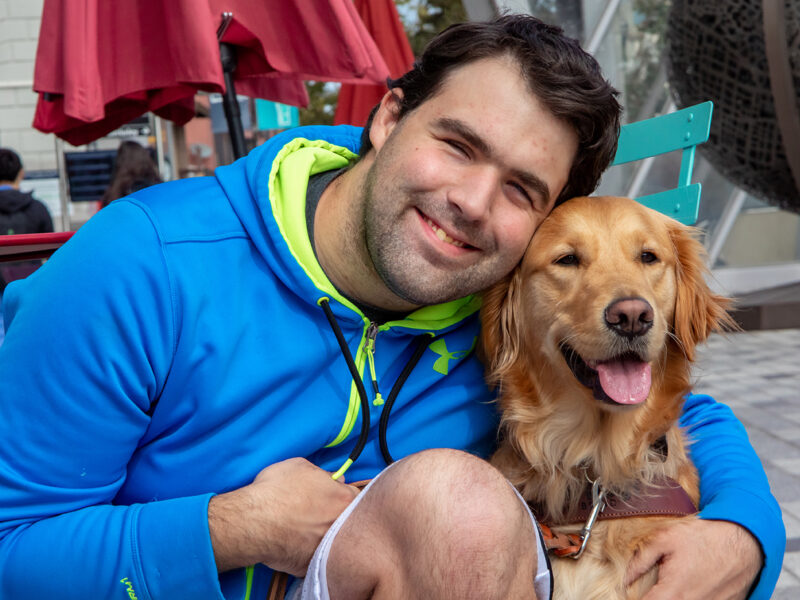 A smiling man in a blue hoodie kneels and embraces his grinning Golden Retriever guide dog.
