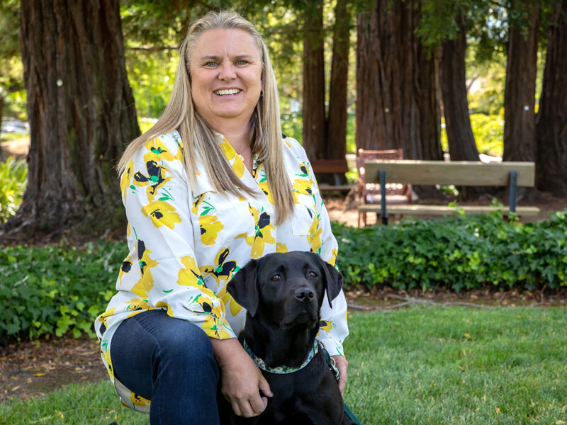 A smiling woman wearing a white shirt with yellow flowers kneels on a lawn next to a black Labrador Retriever