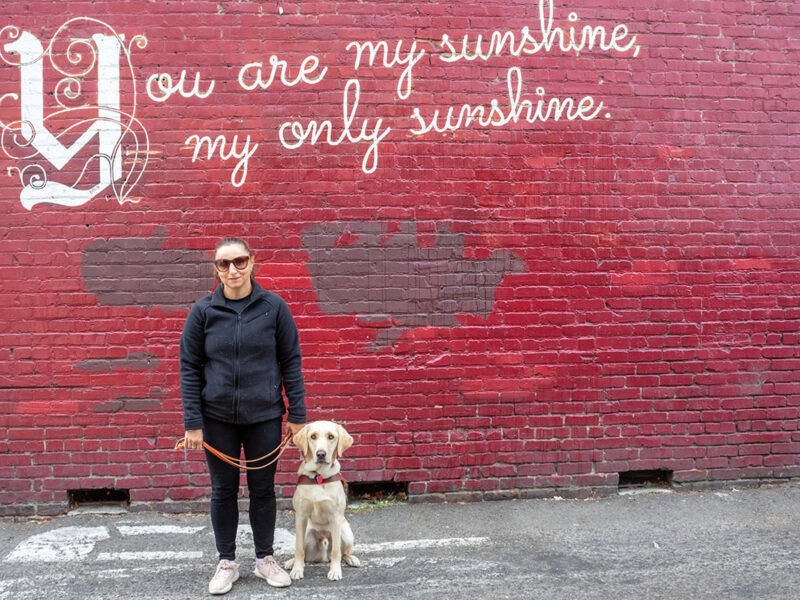 A woman and her guide dog stand against a red brick wall painted with the words "You are my sunshine, my only sunshine."