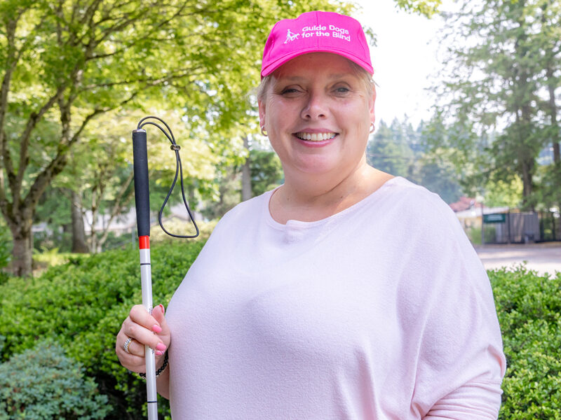 A woman in a pink hat and pink shirt smiles as she poses for a portrait and holds her white cane.