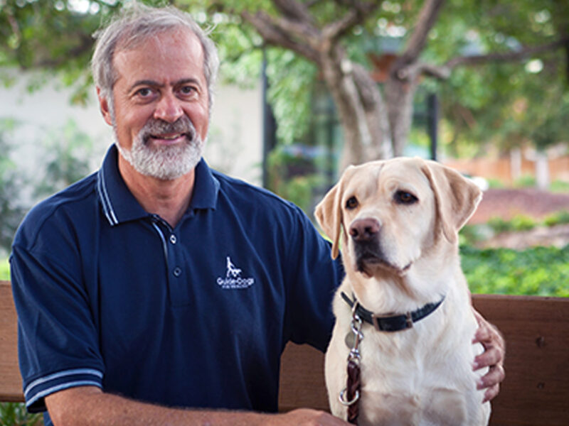 A man sits on a bench with his arm around a yellow Labrador Retriever.